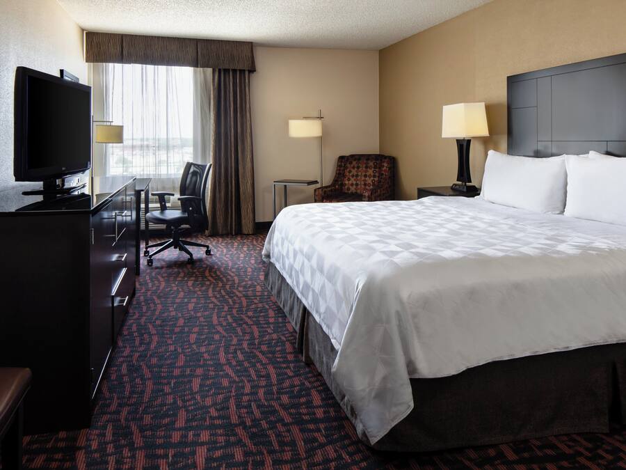 A hotel room with a bed, a desk with a TV, a chair, a lamp, and a window with curtains.