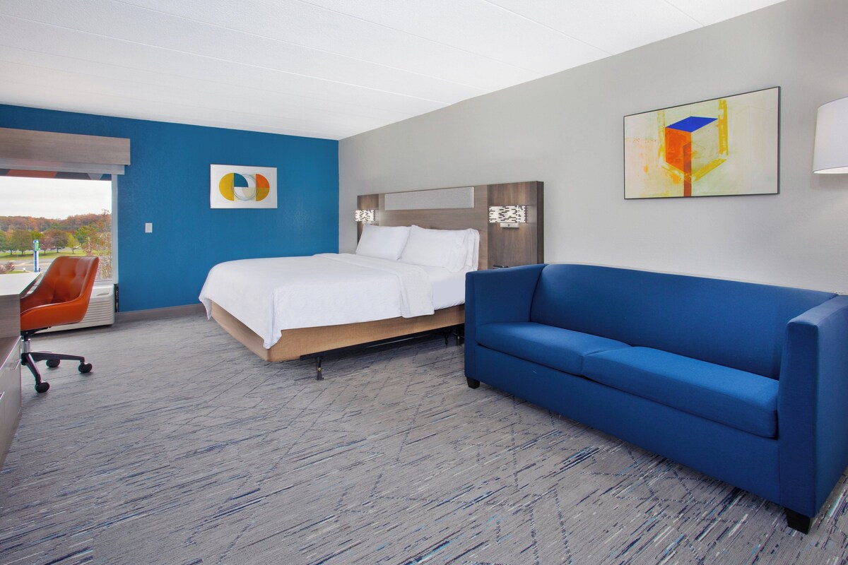 A modern hotel room with a bed, a blue sofa, a desk with a chair, and a large window offering a view of trees.