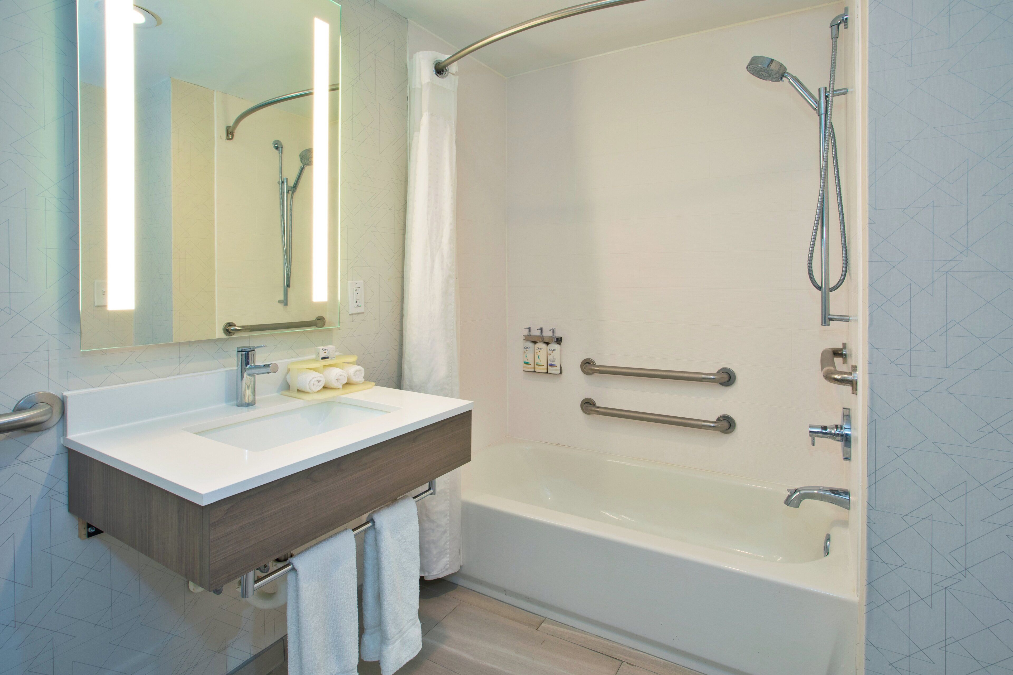 A modern bathroom with a bathtub, a shower, a vanity with a mirror, and handrails.