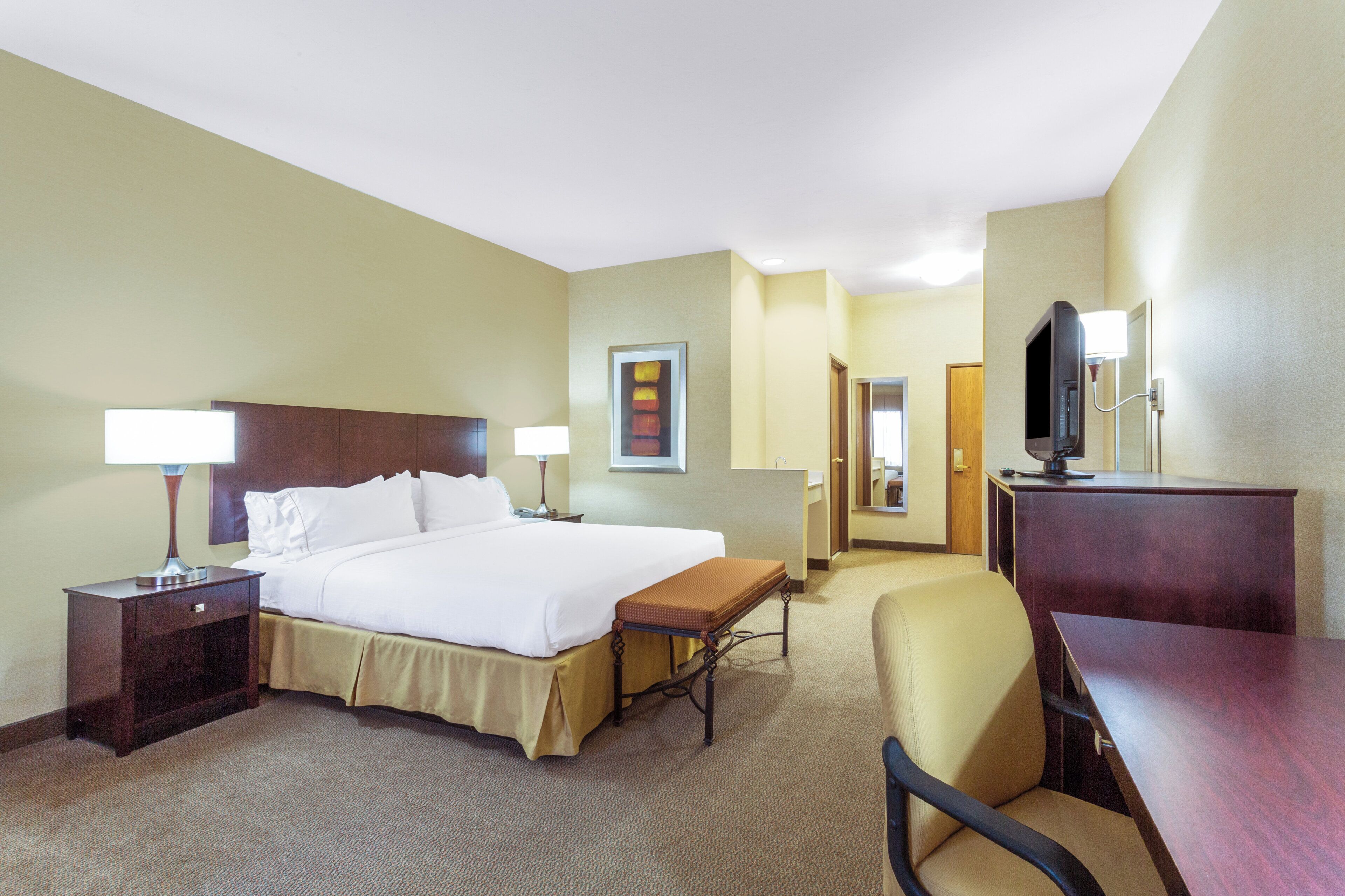 A hotel room with a bed, bedside tables, a desk, a chair, a television, and a bathroom visible through an open door.