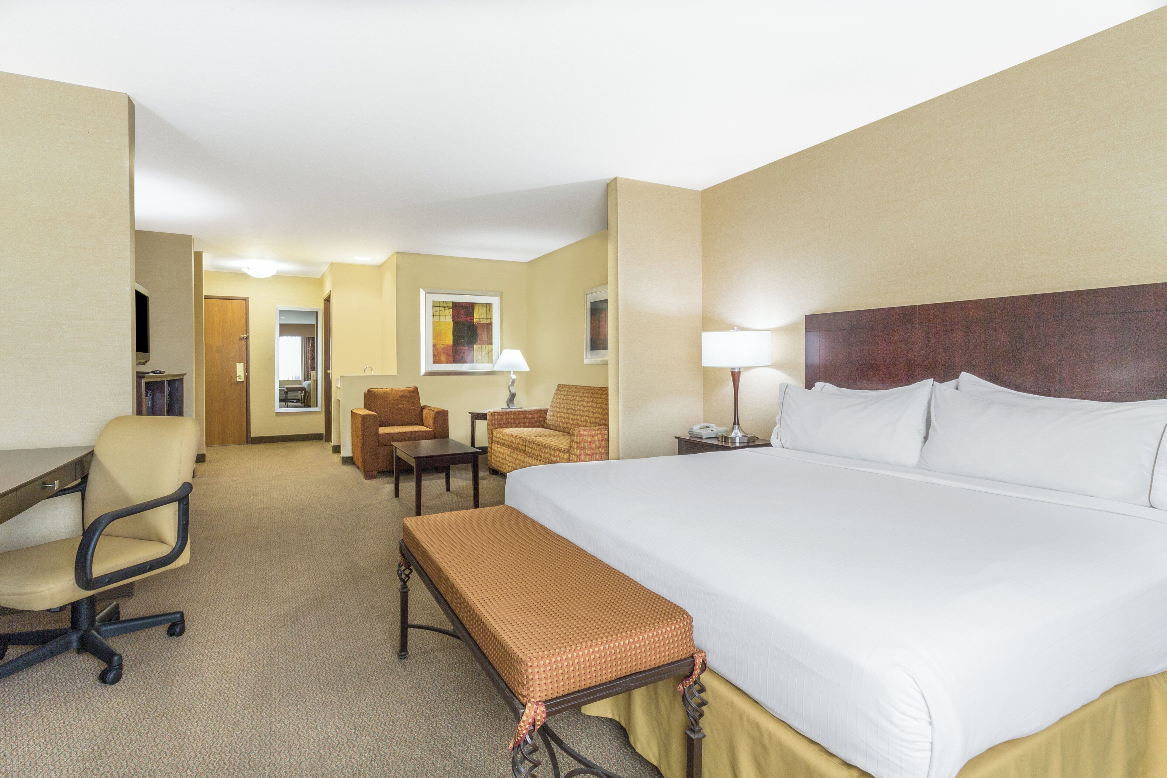A hotel room with a large bed, a desk, a chair, a small table, and a bench.