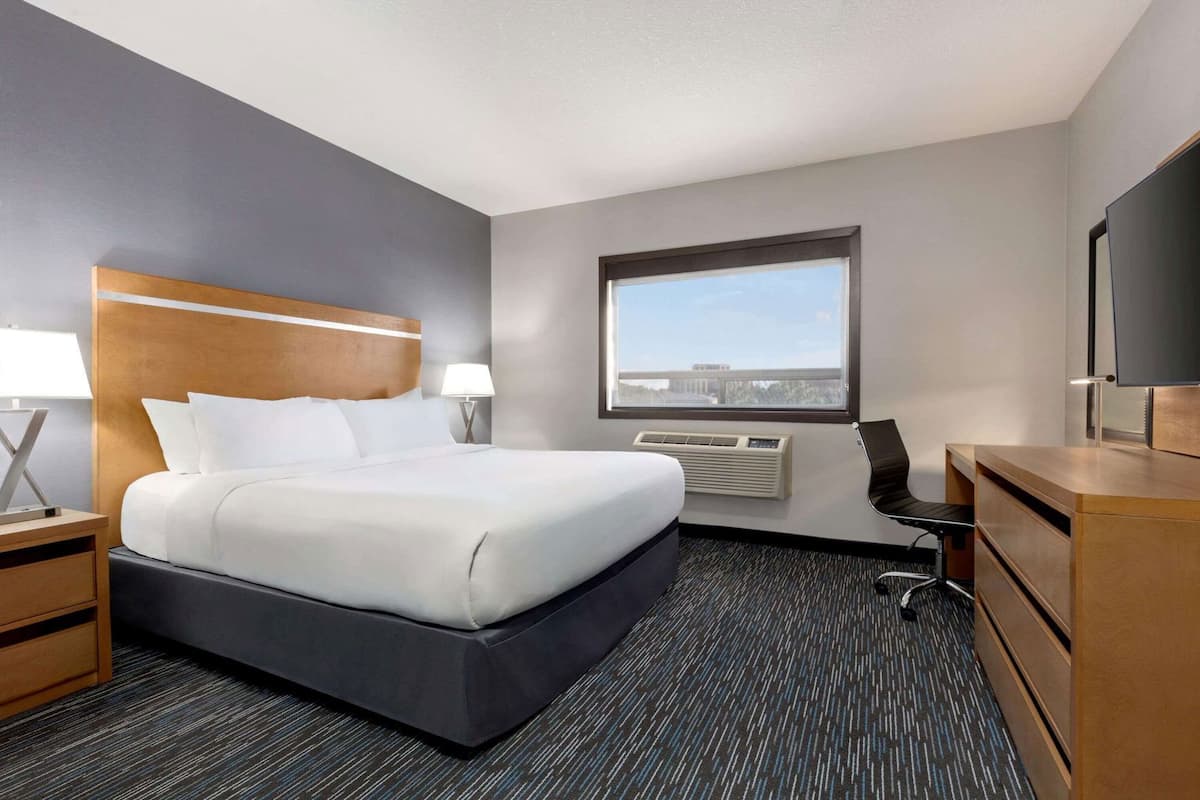A hotel room with a bed, bedside table, lamp, air conditioning unit, window, and a TV.