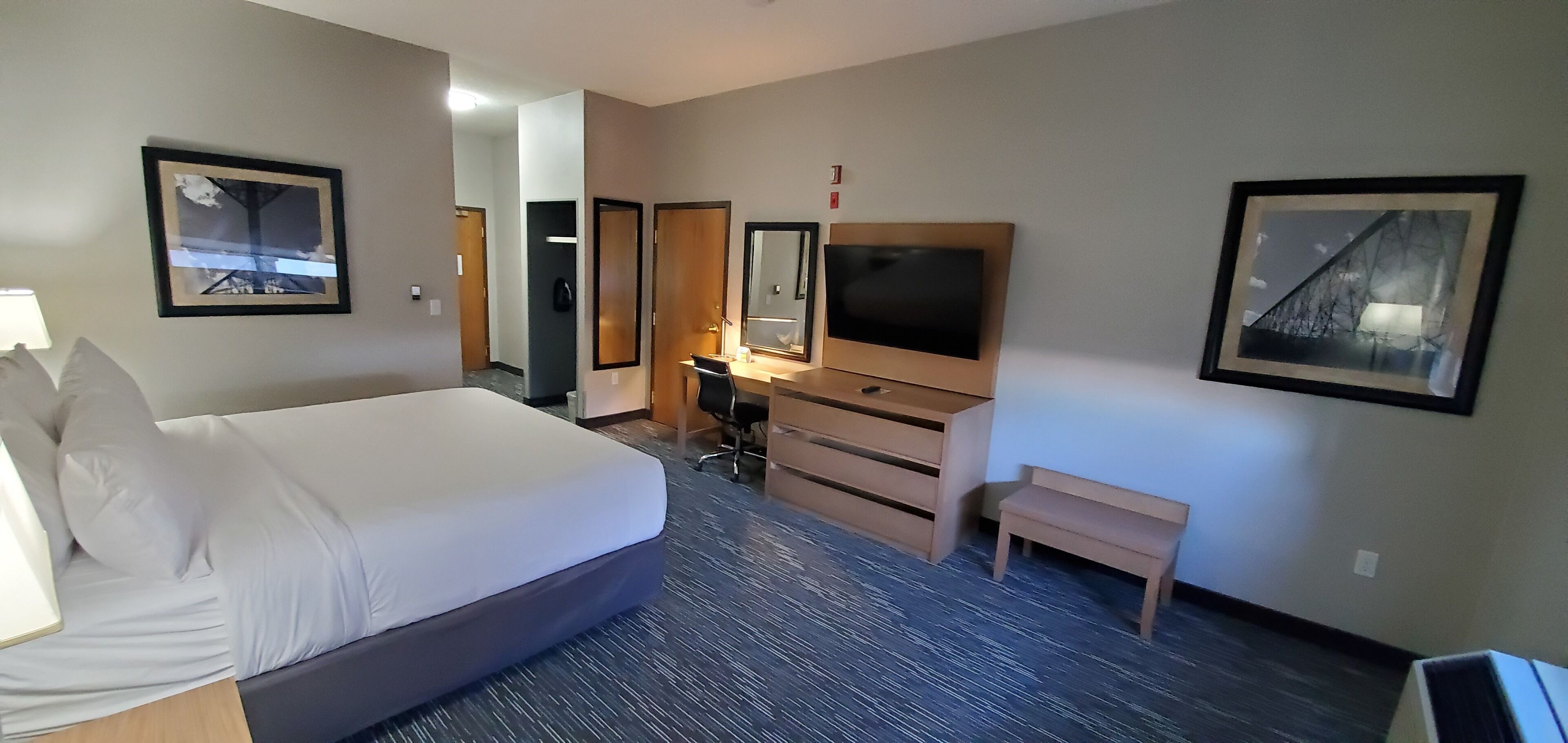 A hotel room with a large bed, a desk with a chair, a TV, a small bench, and framed pictures on the walls.