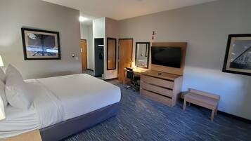 A hotel room with a large bed, a desk with a chair, a TV, a small bench, and framed pictures on the walls.
