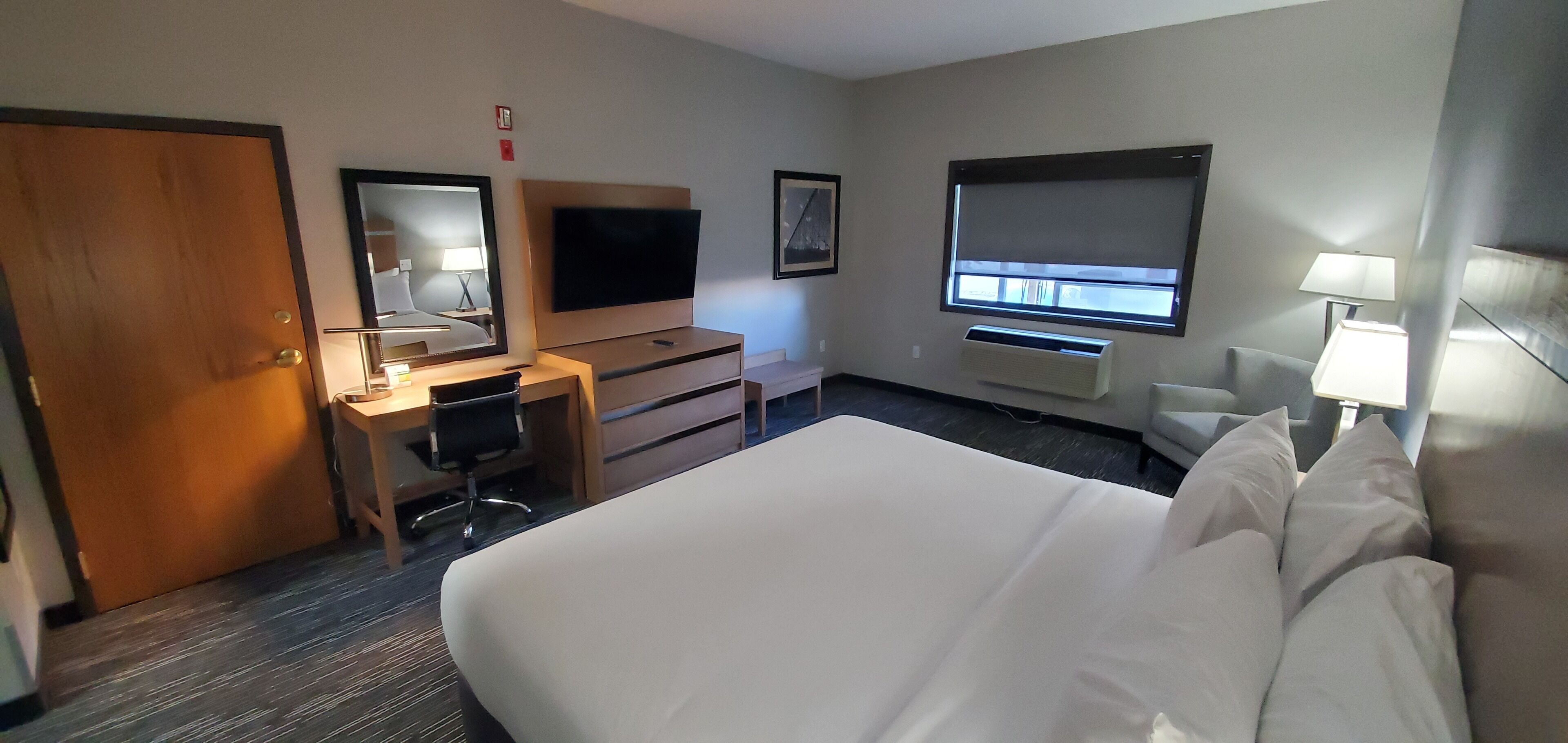 A hotel room with a large bed, a desk with a chair, a television, a window, and a wall-mounted air conditioning unit.