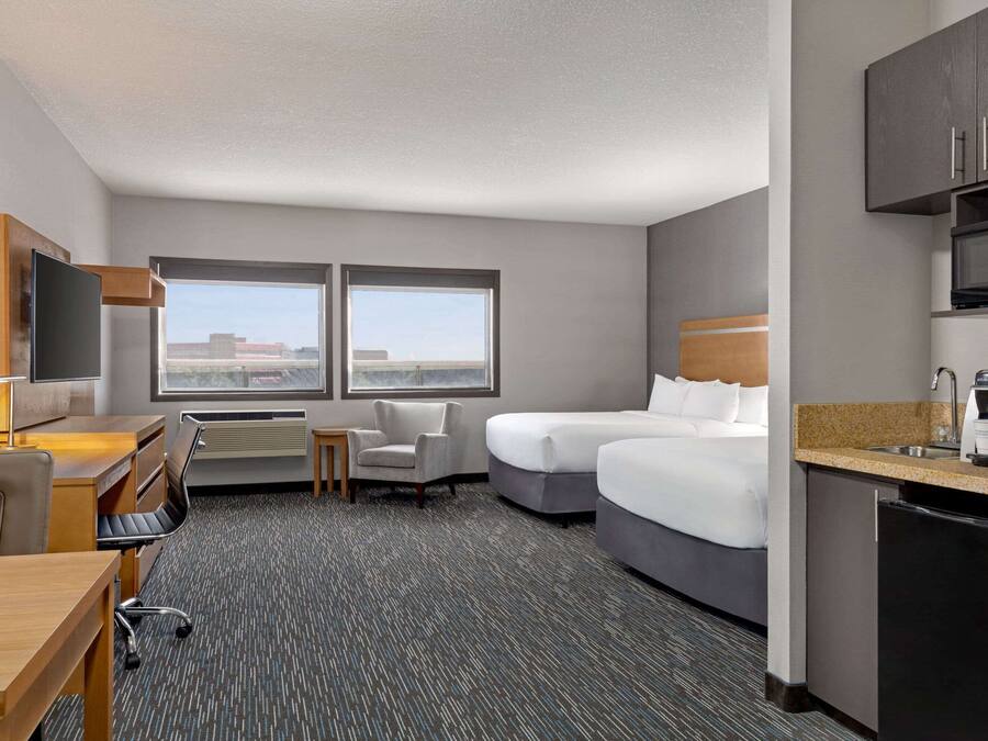 A hotel room with a desk, two beds, a kitchenette, and large windows offering a city view.