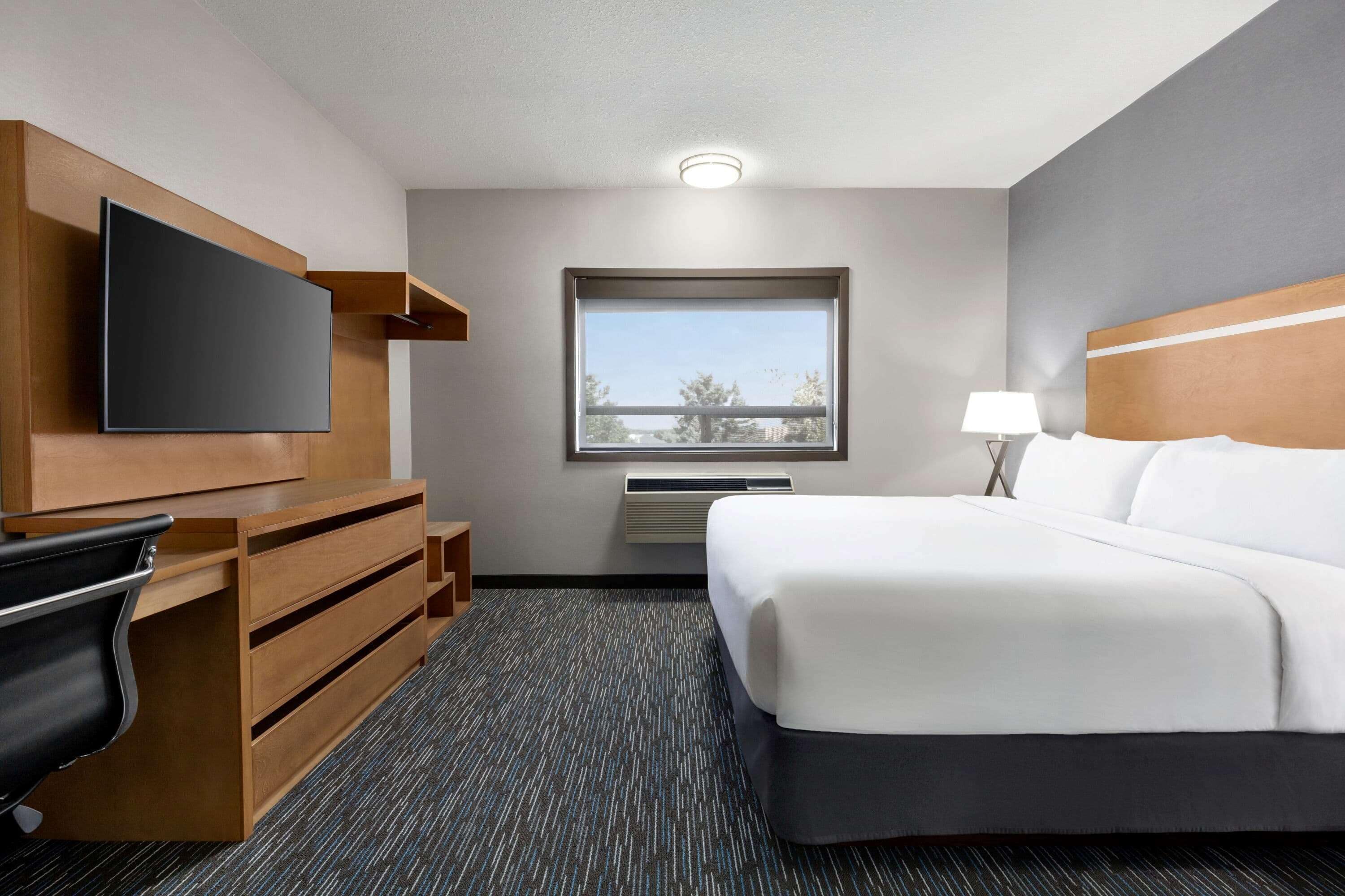 A hotel room with a bed, a flat-screen TV, a desk with drawers, a lamp, and a window with a view.