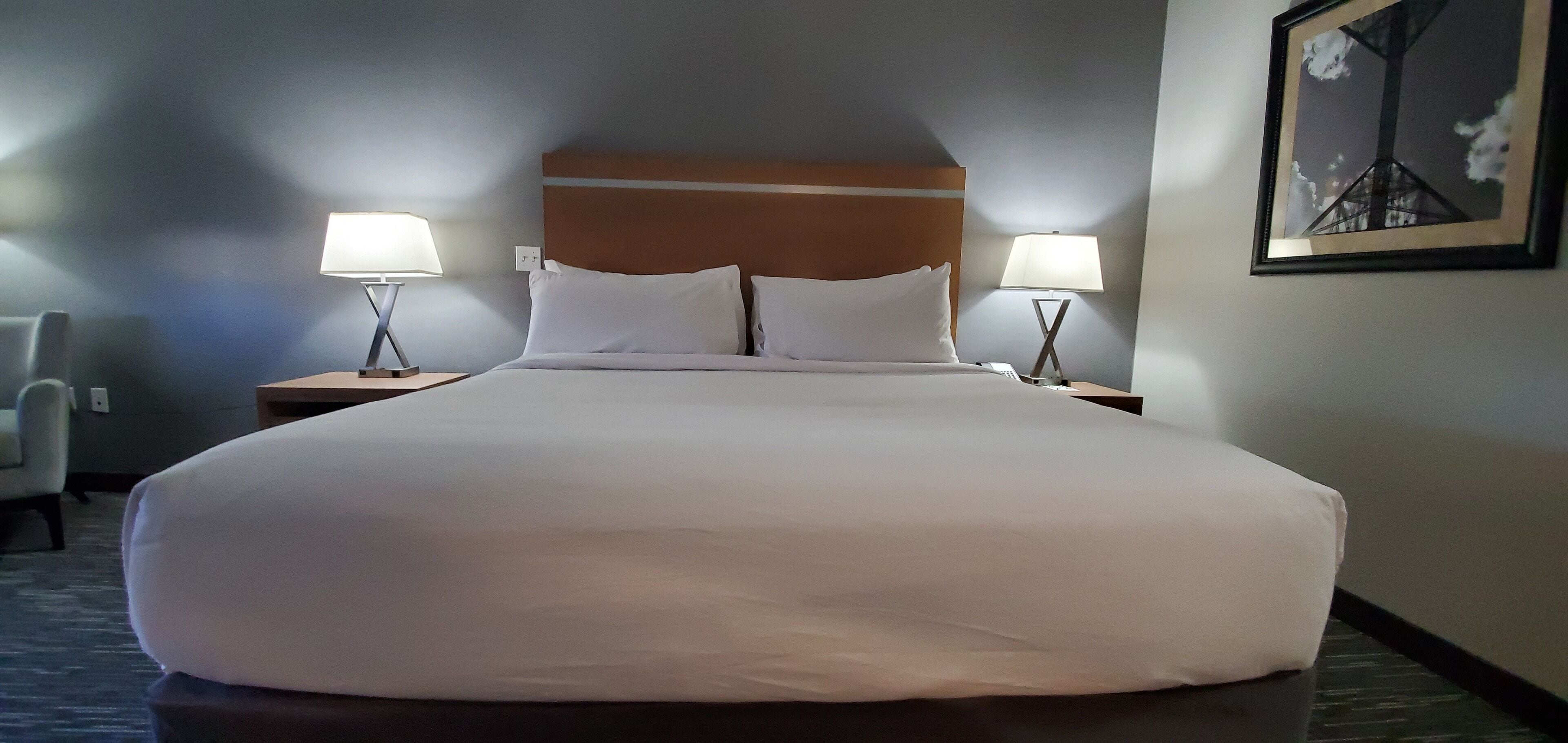 A large bed with white linens, two bedside tables with lamps, a chair, and a framed picture on the wall.
