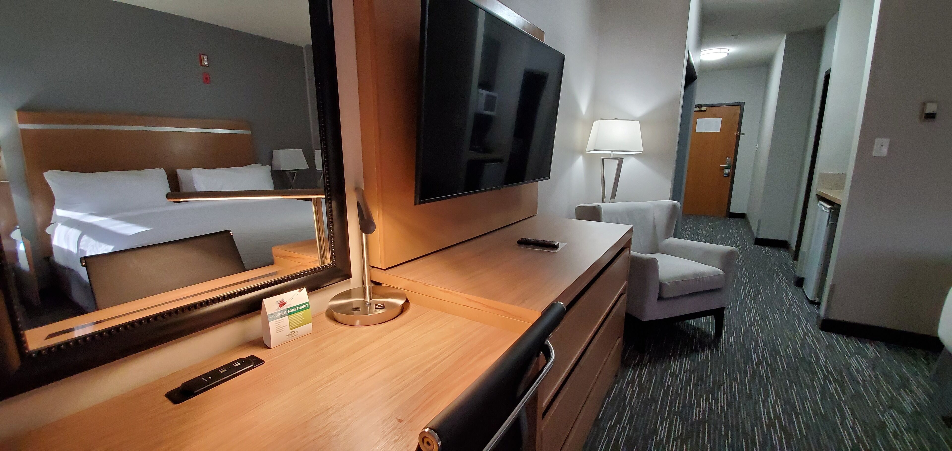 A hotel room with a bed, a TV, a desk with a lamp, a chair, and a carpeted floor.