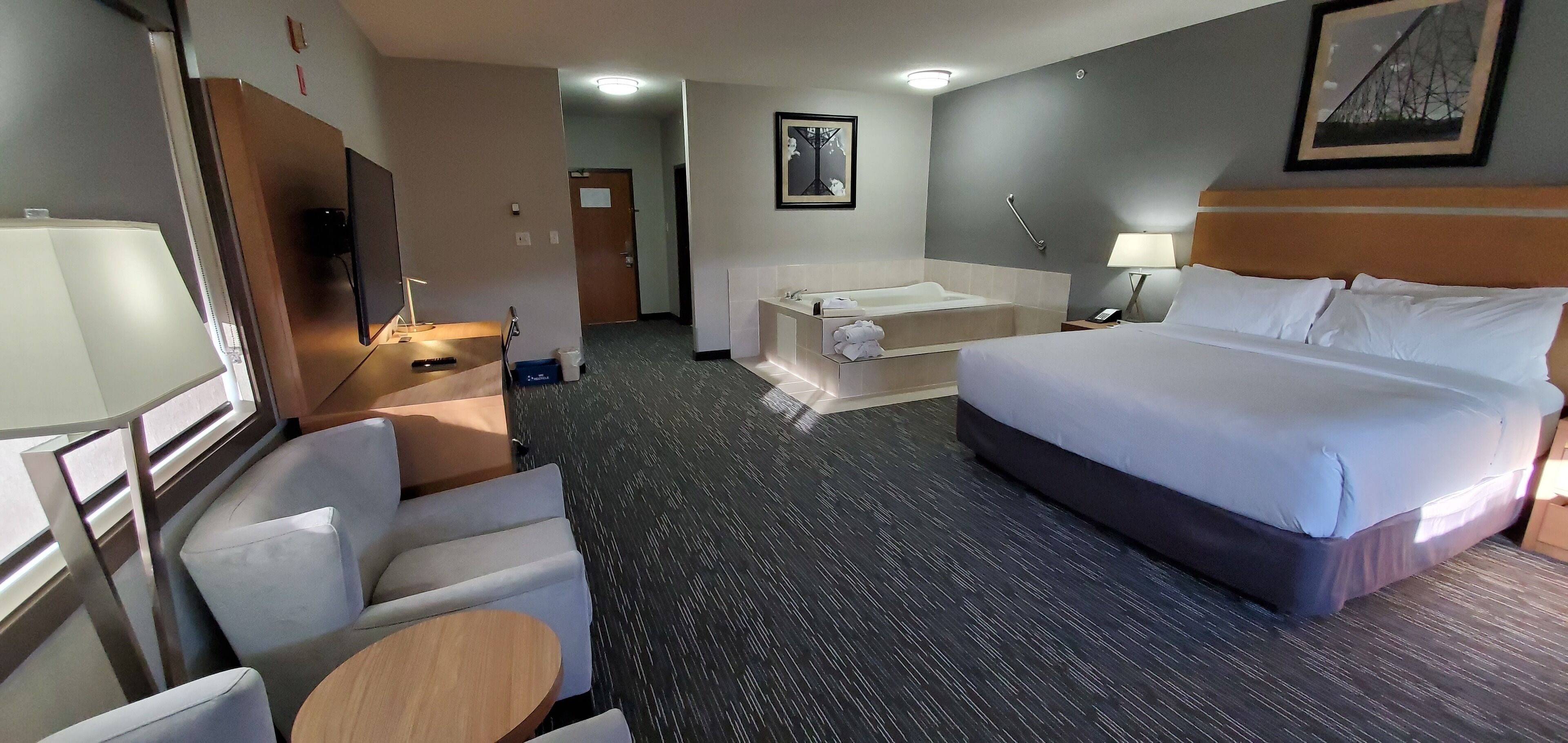 A hotel room with a bed, a desk with a chair, a small table, a bathtub, and a TV.