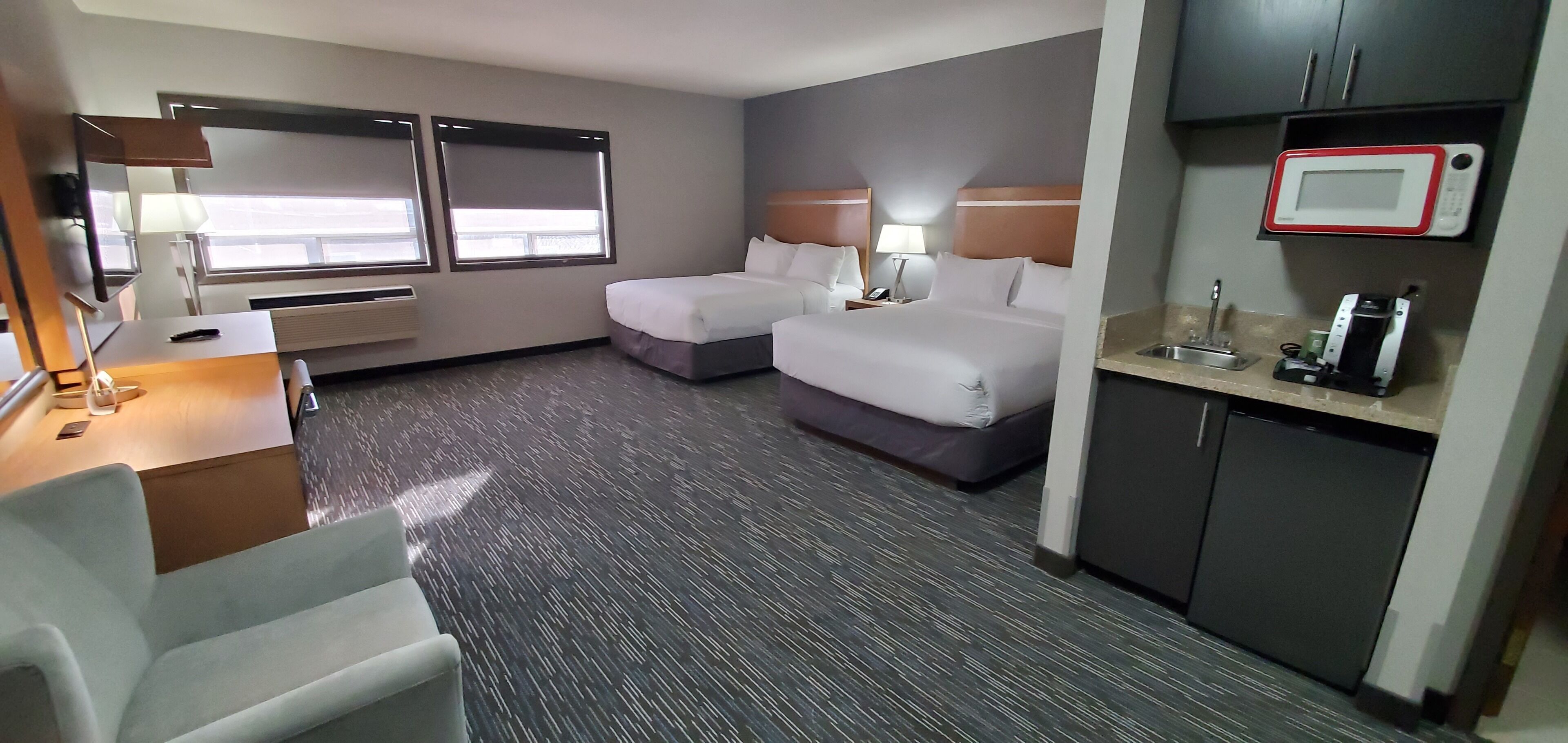 A hotel room with two beds, a desk with a lamp, a small kitchenette, and a window with blinds.