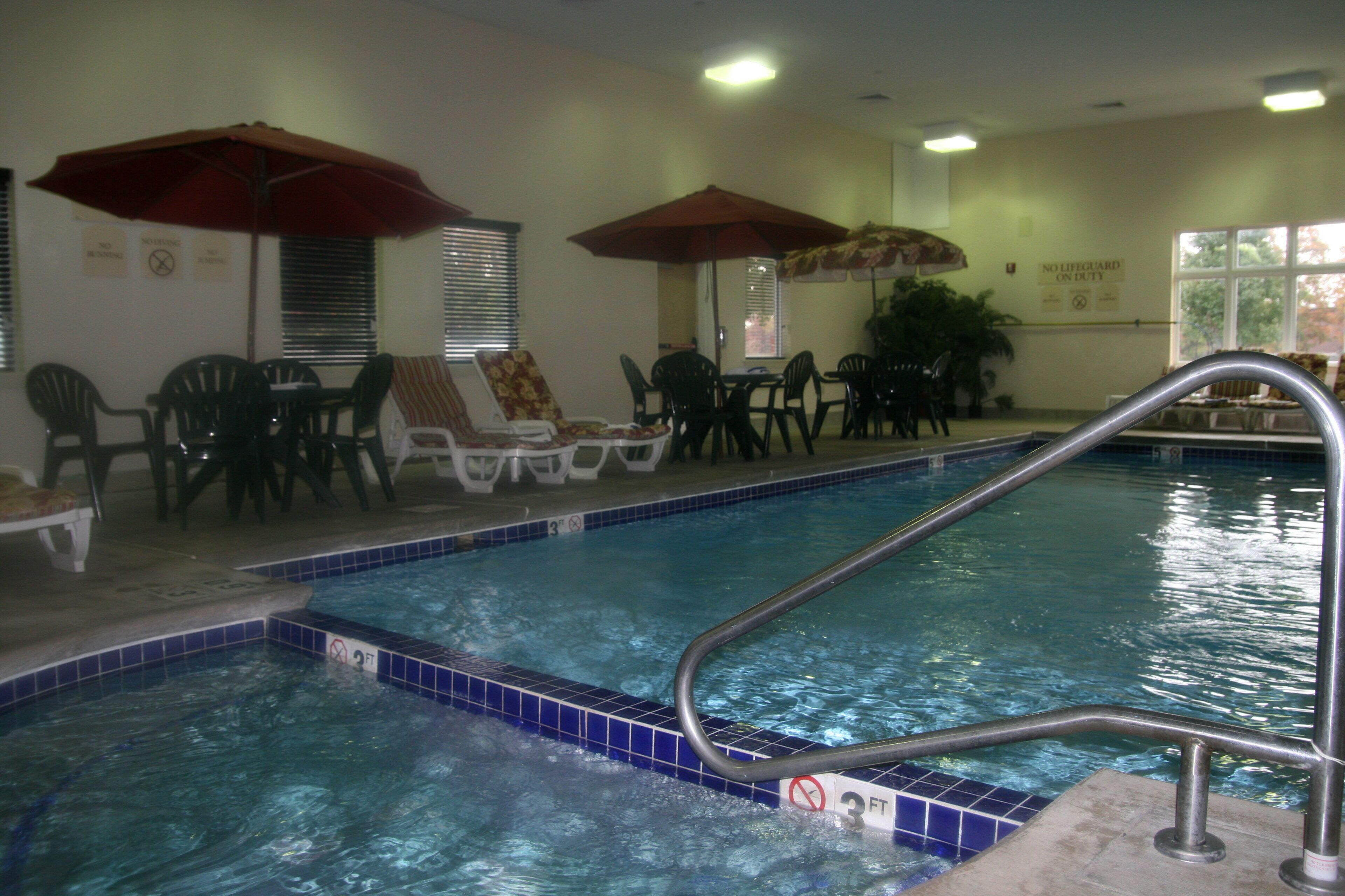 An indoor swimming pool with lounge chairs and umbrellas, a tiled floor, and a no lifeguard on duty sign.