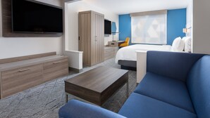 A modern hotel room with a large flat-screen TV, a wooden wardrobe, a desk with a chair, a blue sofa, and a bed with white pillows.