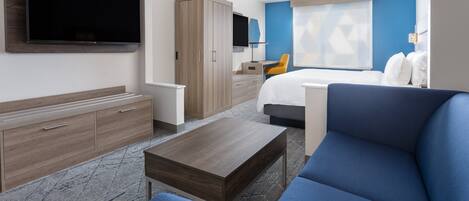 A modern hotel room with a large flat-screen TV, a wooden wardrobe, a desk with a chair, a blue sofa, and a bed with white pillows.