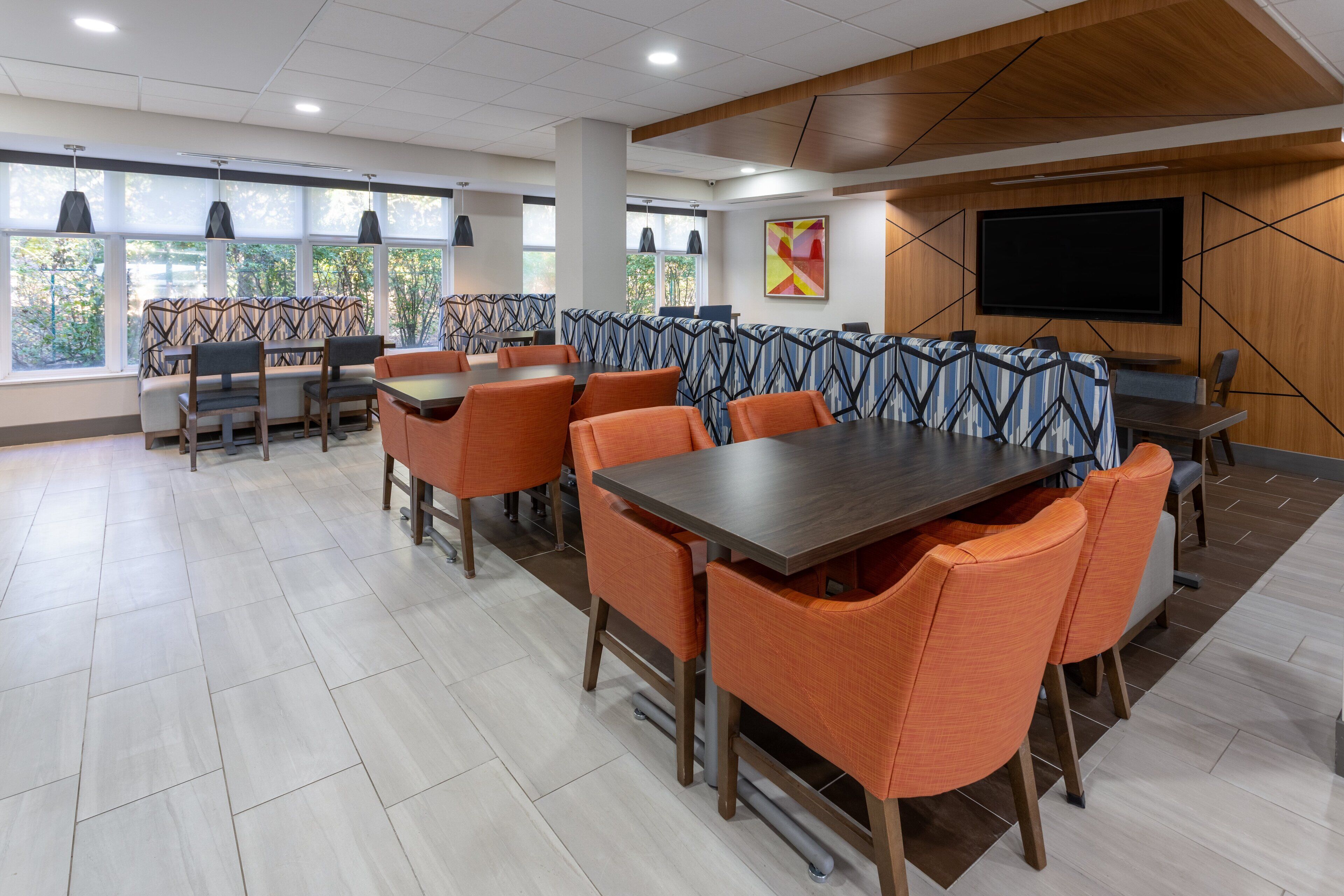 Un comedor moderno con mesas altas y sillas naranjas, ventanas amplias con luz natural y un televisor de pantalla plana en la pared.
