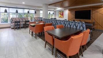 Un comedor moderno con mesas altas y sillas naranjas, ventanas amplias con luz natural y un televisor de pantalla plana en la pared.