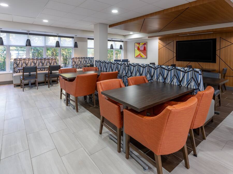 Un comedor moderno con mesas altas y sillas naranjas, ventanas amplias con luz natural y un televisor de pantalla plana en la pared.