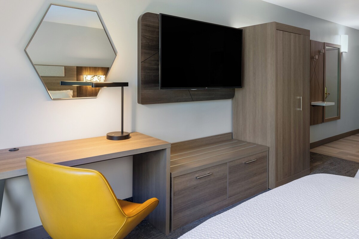 Ein modernes Hotelzimmer mit einem flachbildfernseher an der Wand, einem Holztisch, einem gelben Sessel und einem großen Kleiderschrank.