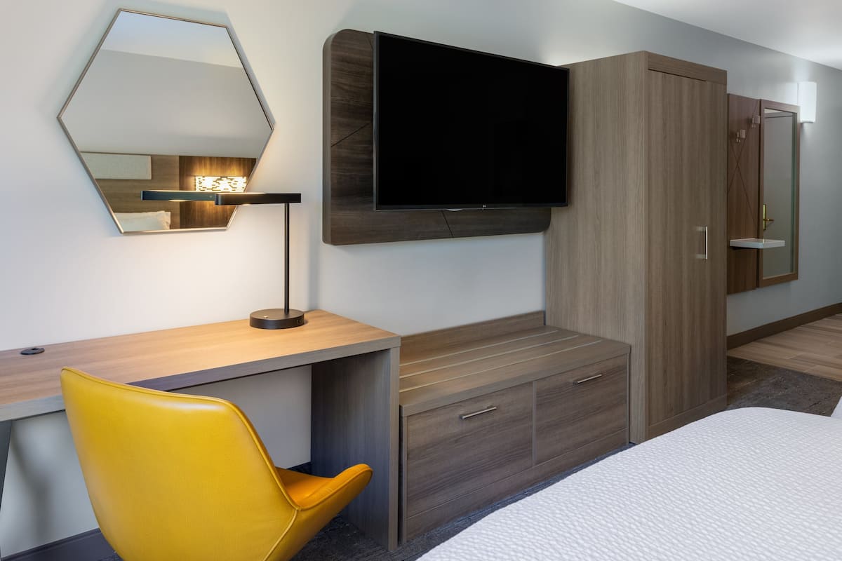 Habitación de hotel moderna con televisor de pantalla plana en la pared, un escritorio de madera, una silla amarilla y un armario grande.