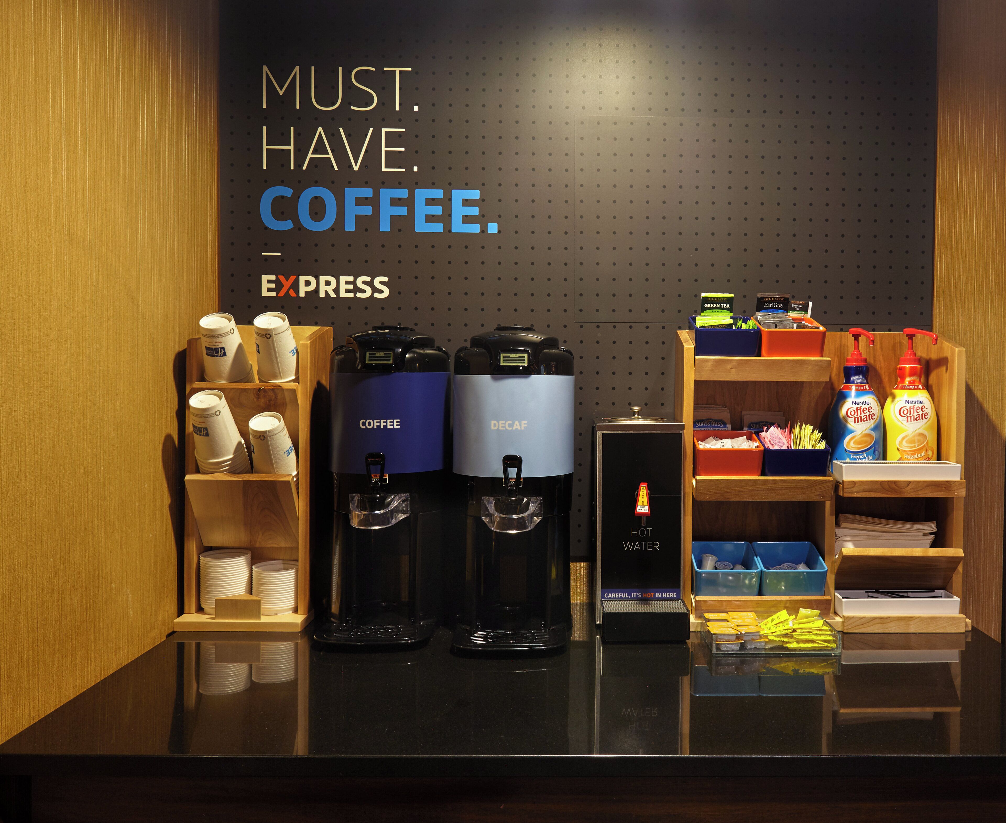 A coffee station with two coffee machines, a hot water dispenser, and a variety of coffee products and condiments.