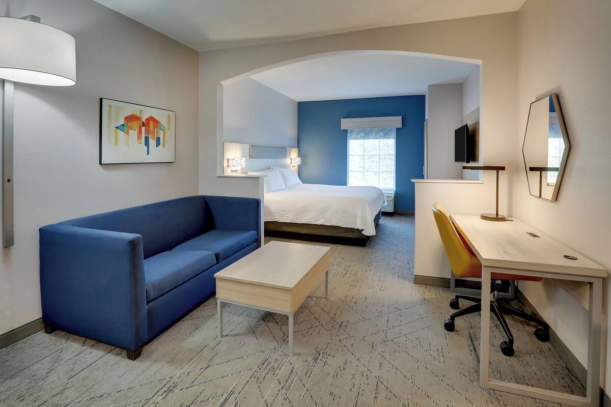 A hotel room with a bed, a desk, a chair, a sofa, a lamp, and a painting.