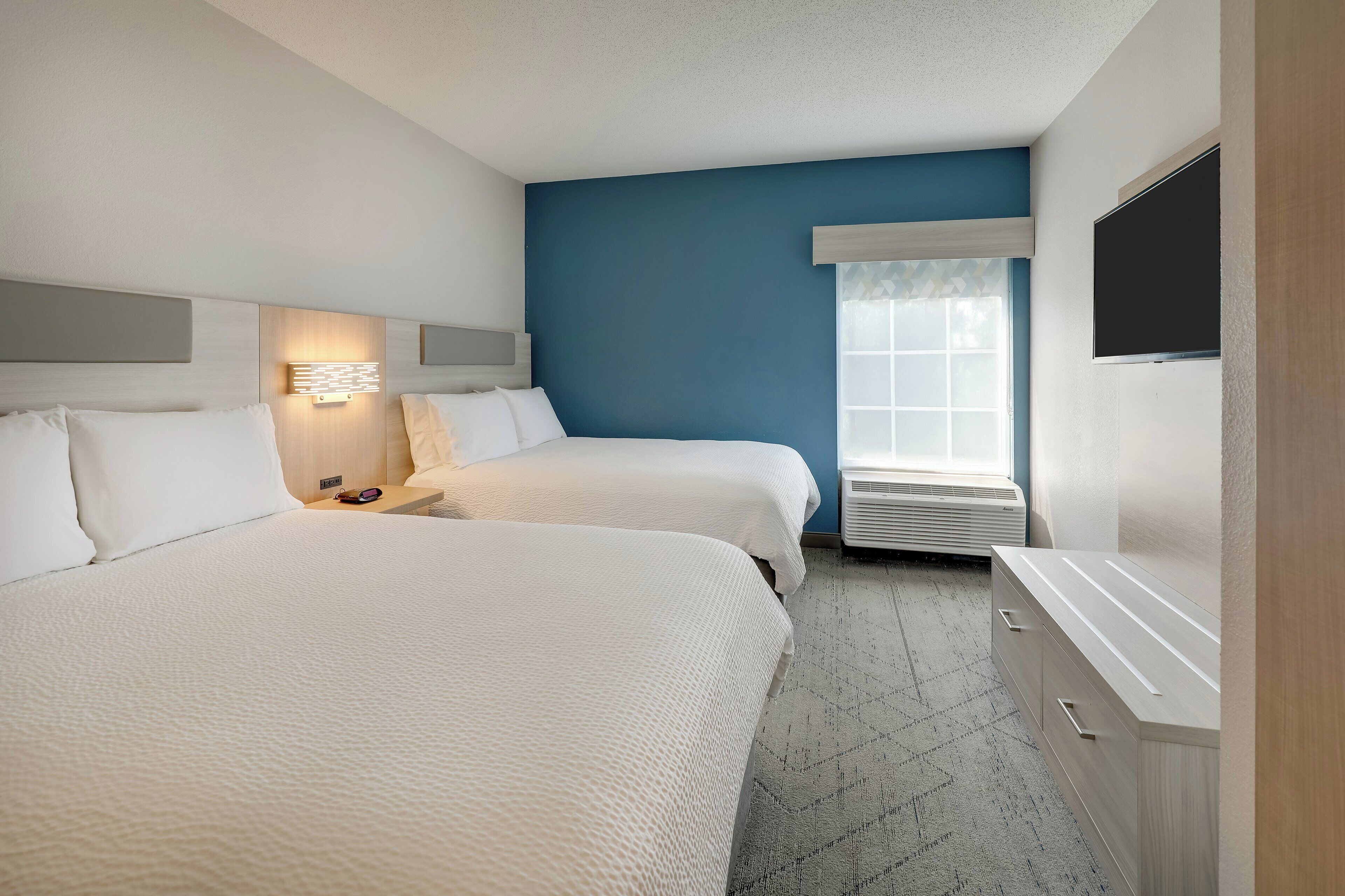 A hotel room with two beds, a bedside table with a lamp, a large window with blinds, and a flat-screen TV mounted on the wall.