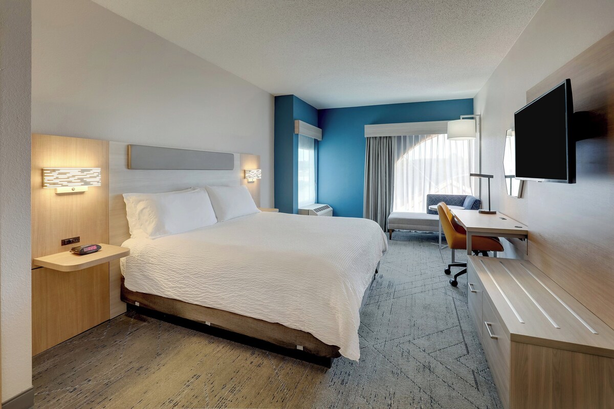 A hotel room with a bed, bedside tables, a desk, a chair, a flat-screen TV, and a window with curtains.