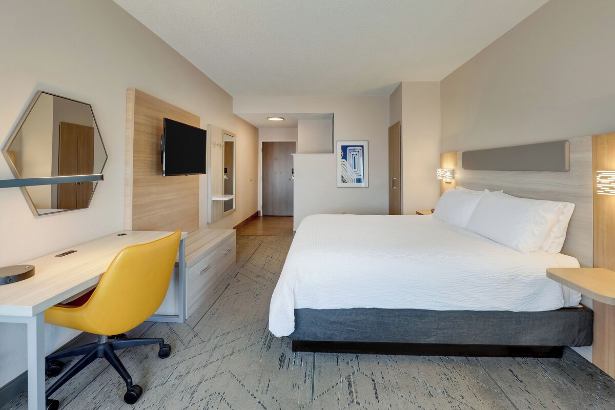 A hotel room with a bed, a desk with a chair, a TV, and a painting on the wall.