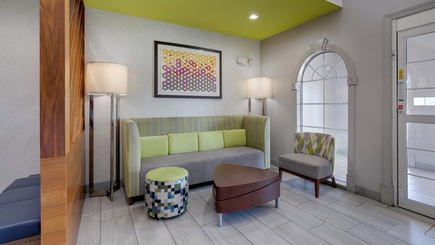 A modern hotel lobby with a green ceiling, a grey sofa, a patterned ottoman, a wooden bench, and a chair with a geometric throw. - Holiday Inn Express & Suites Orangeburg by IHG (Orangeburg)