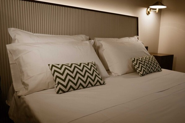 A neatly made bed with white linens and two patterned pillows, a wall-mounted light fixture, and a vertical panel design on the headboard.