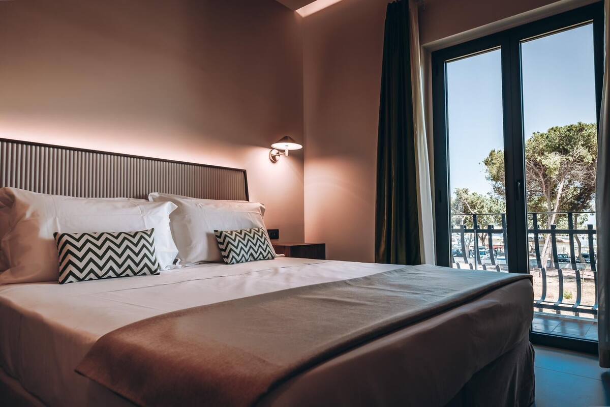 Habitación de hotel con una cama grande, dos almohadas, una mesita de noche, una lámpara de pared y vistas al exterior a través de un ventanal.
