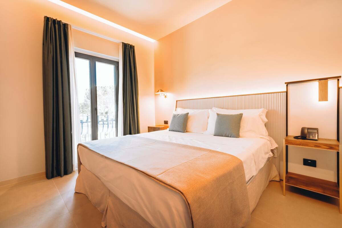 Habitación de hotel con una cama grande, una mesita de noche y un ventanal con cortinas.