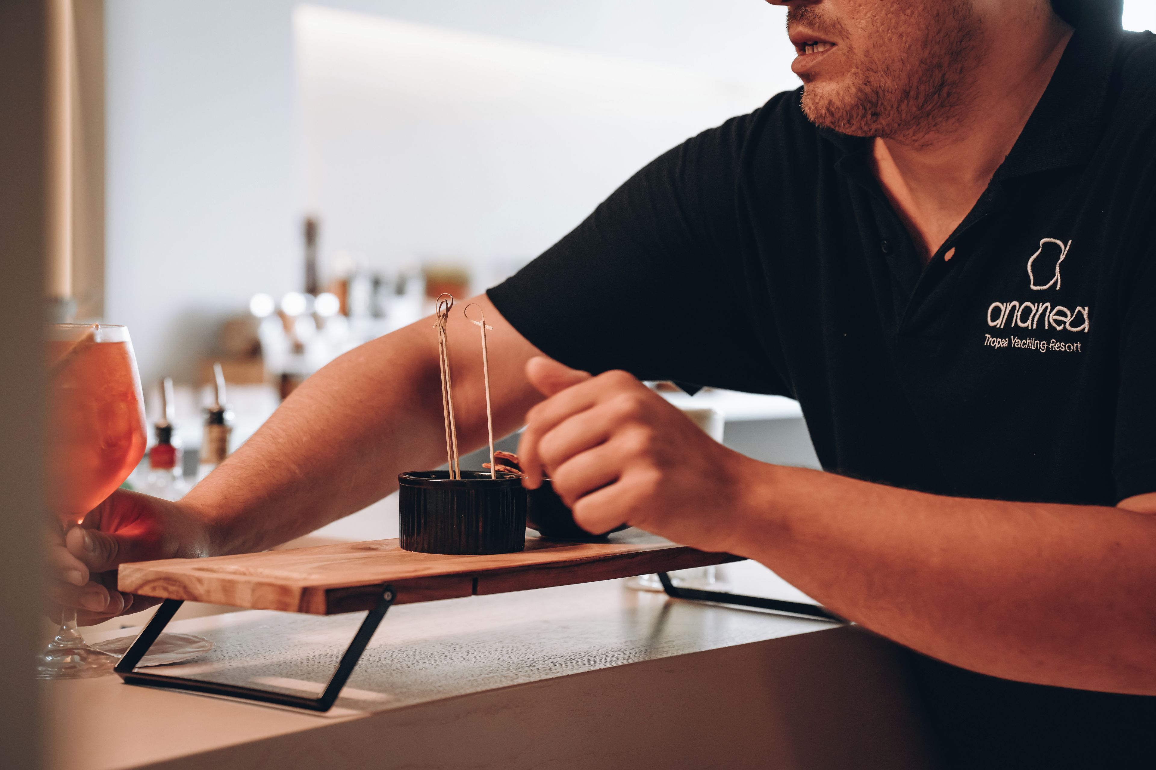 Une personne assise au bar, avec un verre à la main et un plateau rempli de brochettes.