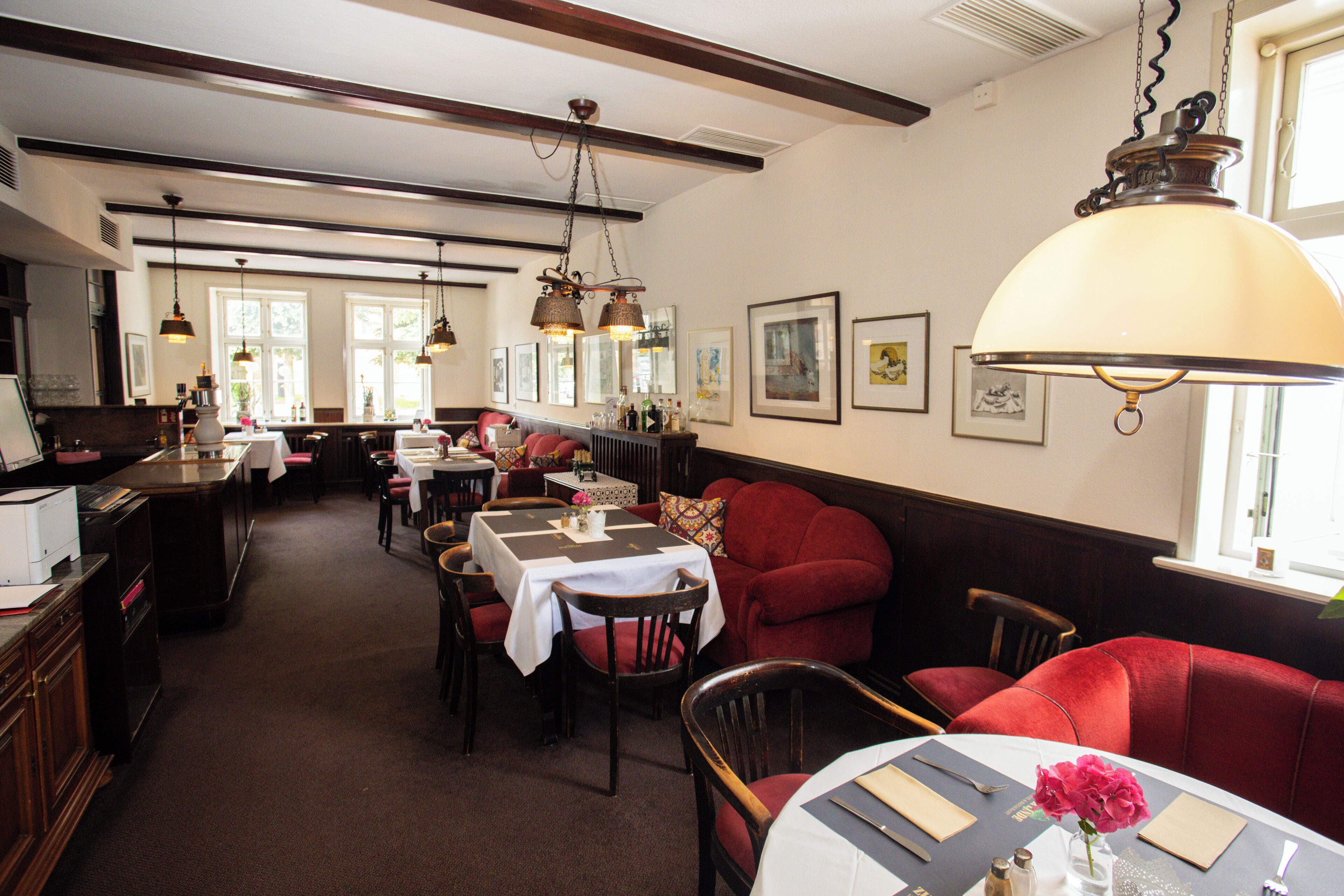 A restaurant with wooden beams, hanging lamps, and red upholstered seating. Tables are set with white tablecloths and napkins.