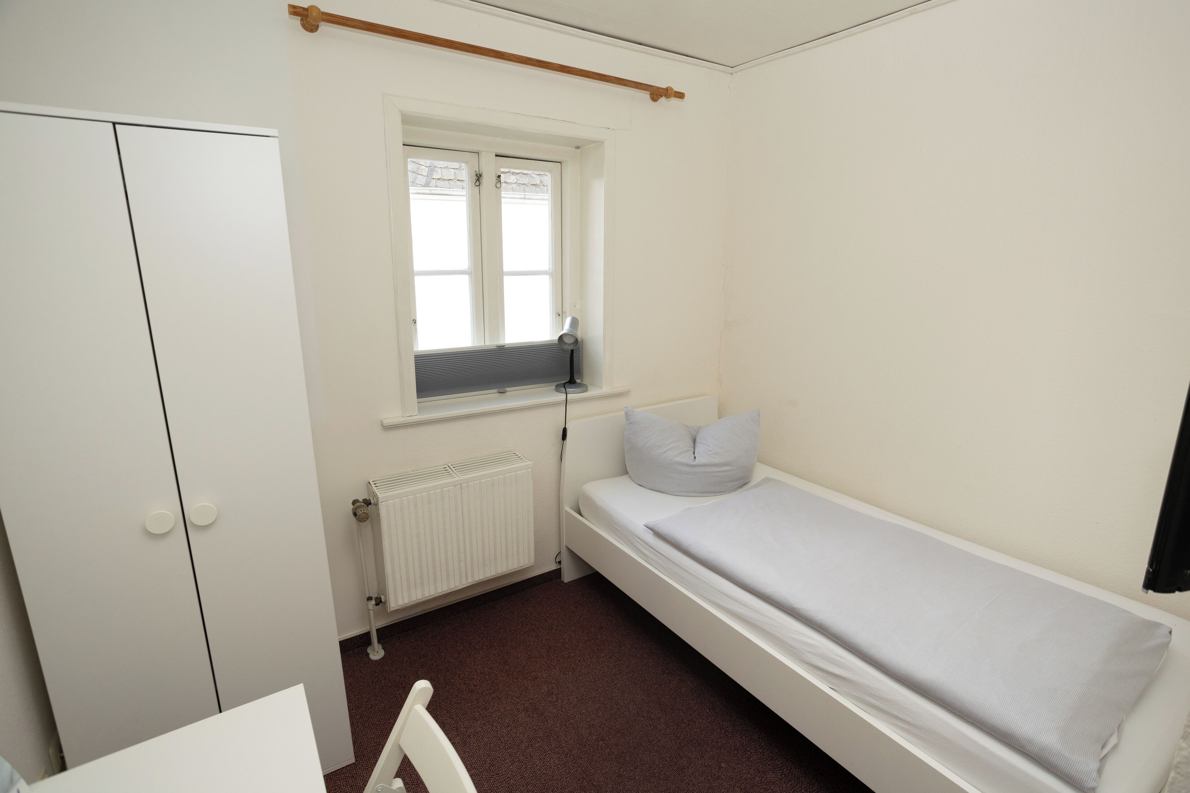 Una habitación pequeña y limpia con una cama individual, un radiador, una ventana con lámpara de escritorio y un armario.