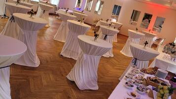 Un salón de banquetes con mesas redondas cubiertas con manteles blancos, suelo de madera y una mesa de bufé con diversos platos.