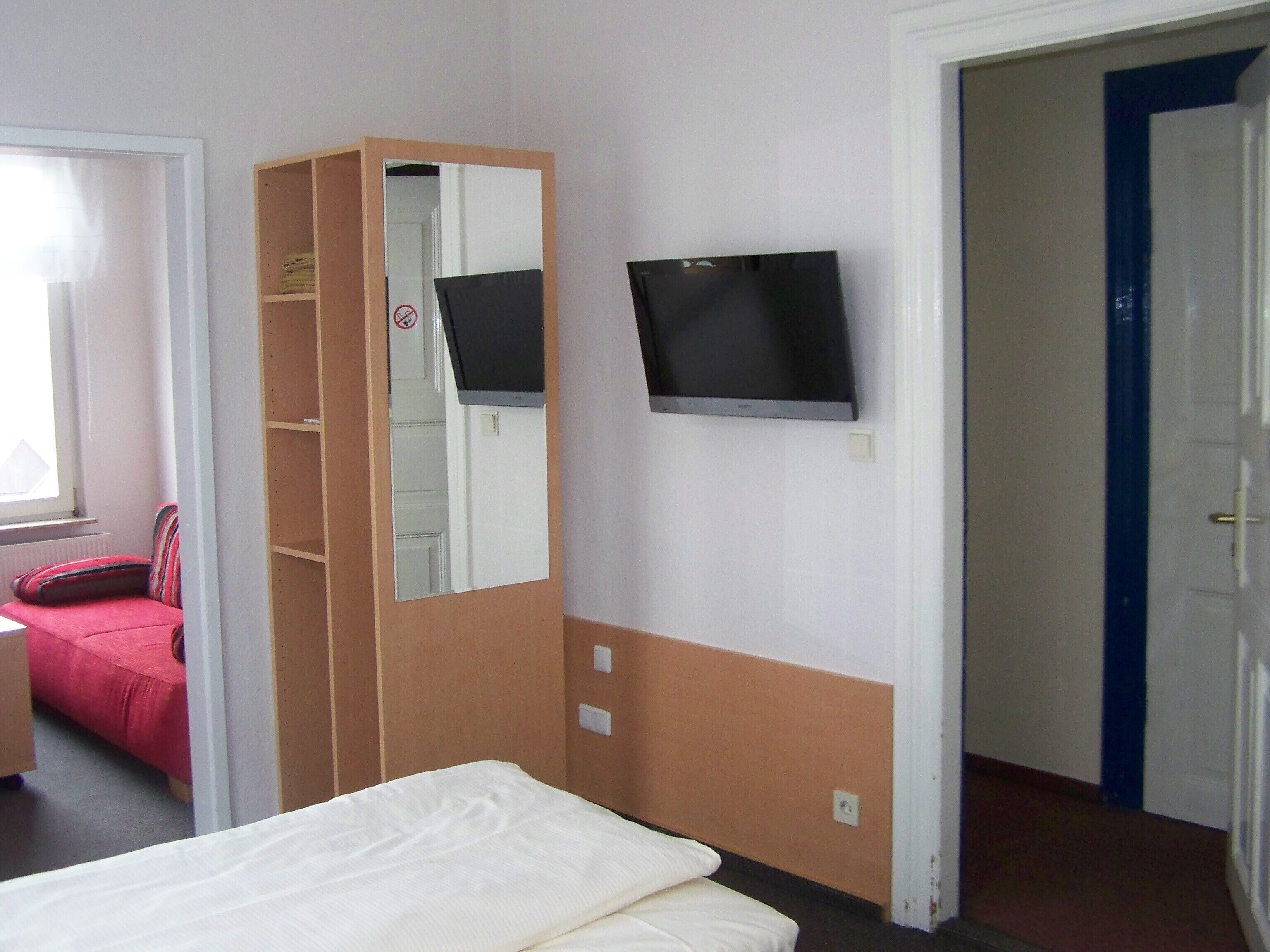 A hotel room with a bed, a television mounted on the wall, a wardrobe, and a door leading to another room.