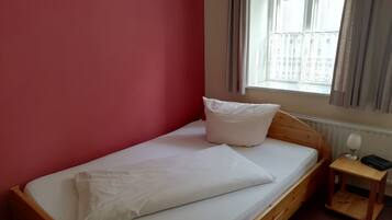 A single bed with white bedding and pillows, a wooden headboard, a small bedside table with a lamp and phone, and a window with curtains.