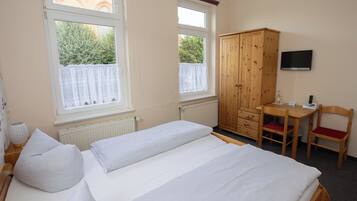 A bedroom with a bed, a wooden wardrobe, a desk with a chair, and a television on the wall.