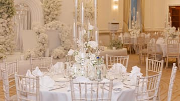 A banquet hall with round tables set for dining, white floral arrangements, and tall candle holders.