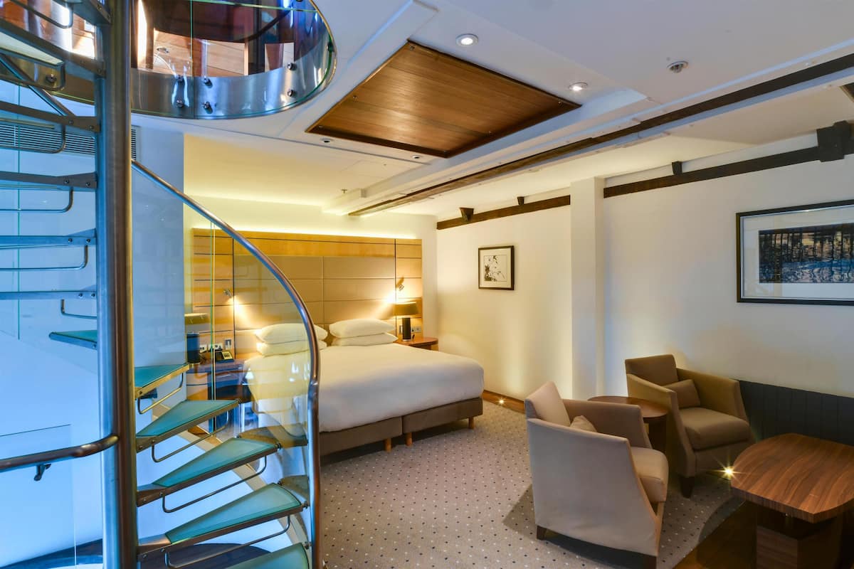 A hotel room with a bed, two chairs, a small table, a staircase, and a wall-mounted picture.