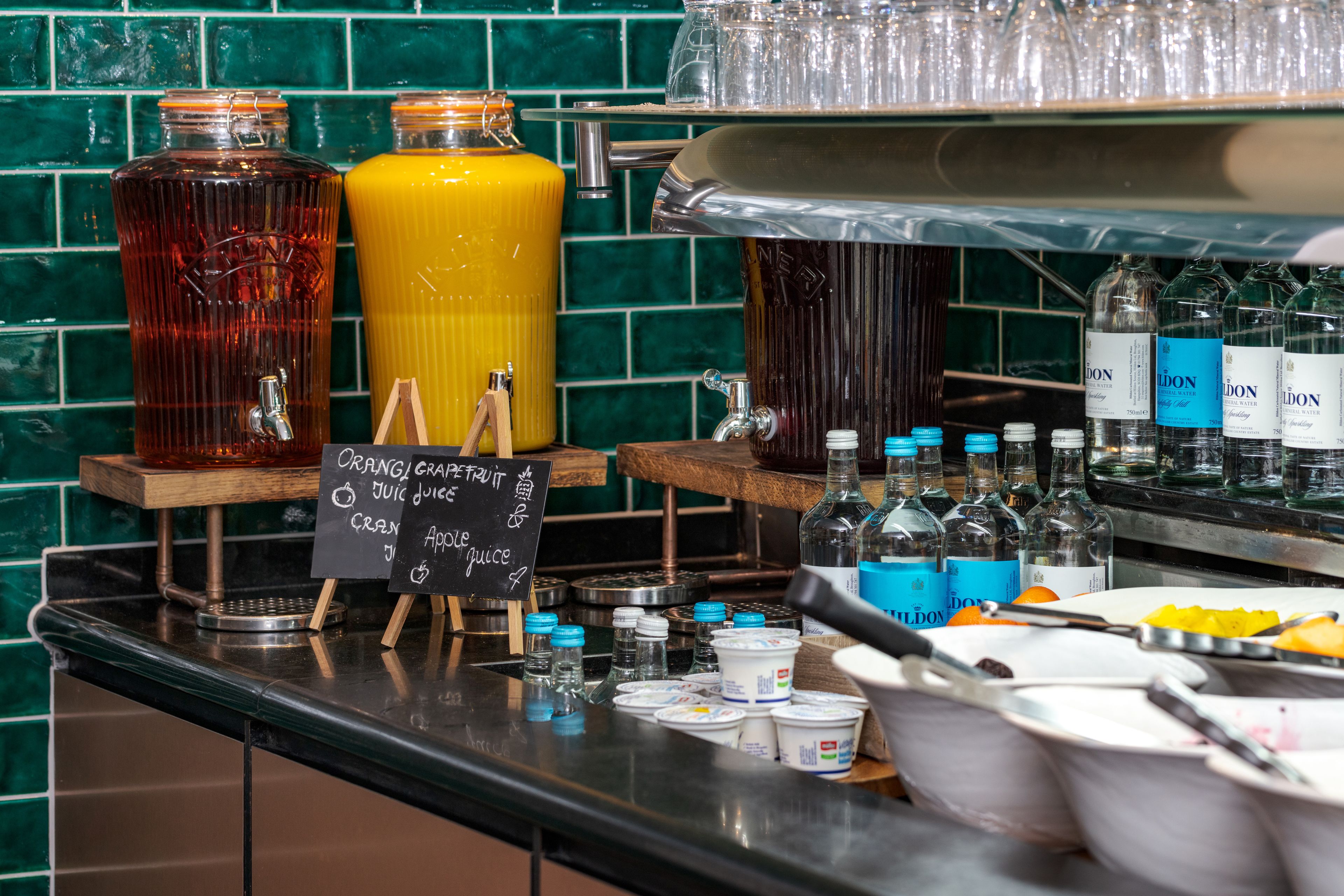 A beverage station with large containers of orange and apple juice, a selection of bottled water, and a chalkboard menu listing juice options.
