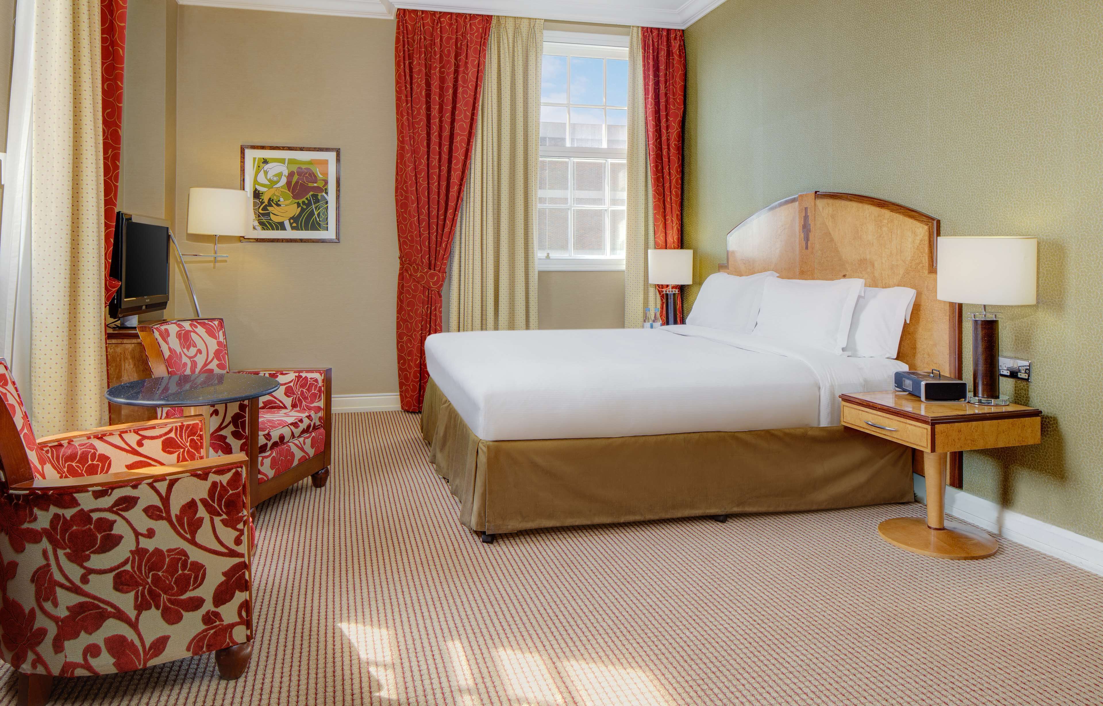 A hotel room with a large bed, two bedside tables with lamps, a small round table, a sofa with floral upholstery, and a television on a stand.