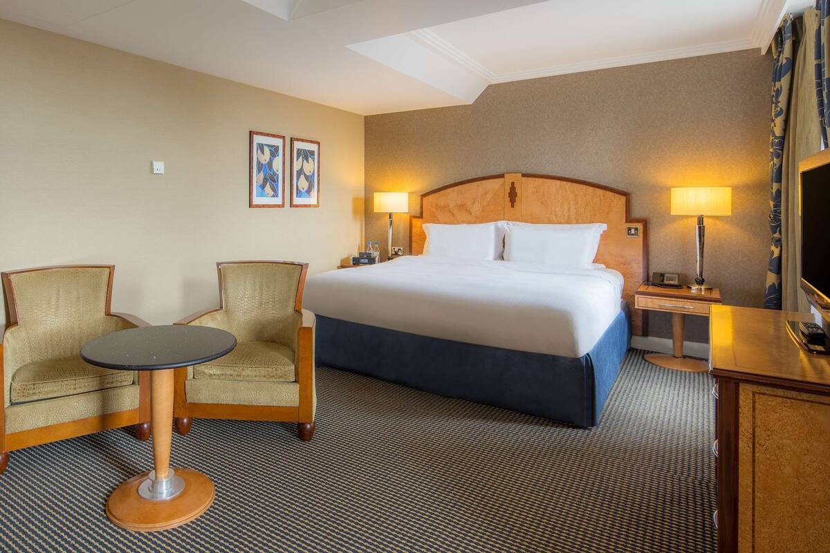 A hotel room with a bed, two armchairs, a small table, a desk with a lamp, and a television.