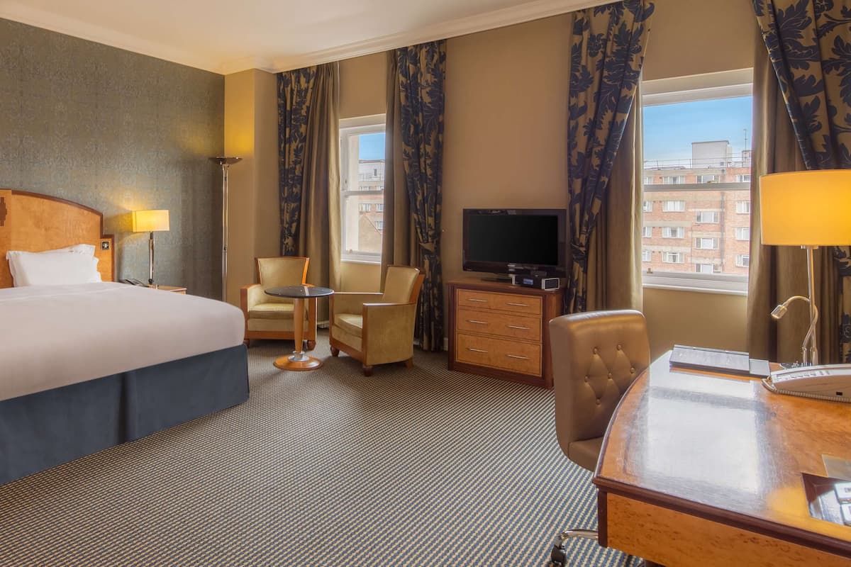 A hotel room with a large bed, a desk, a chair, a TV, and a view of buildings through the window.
