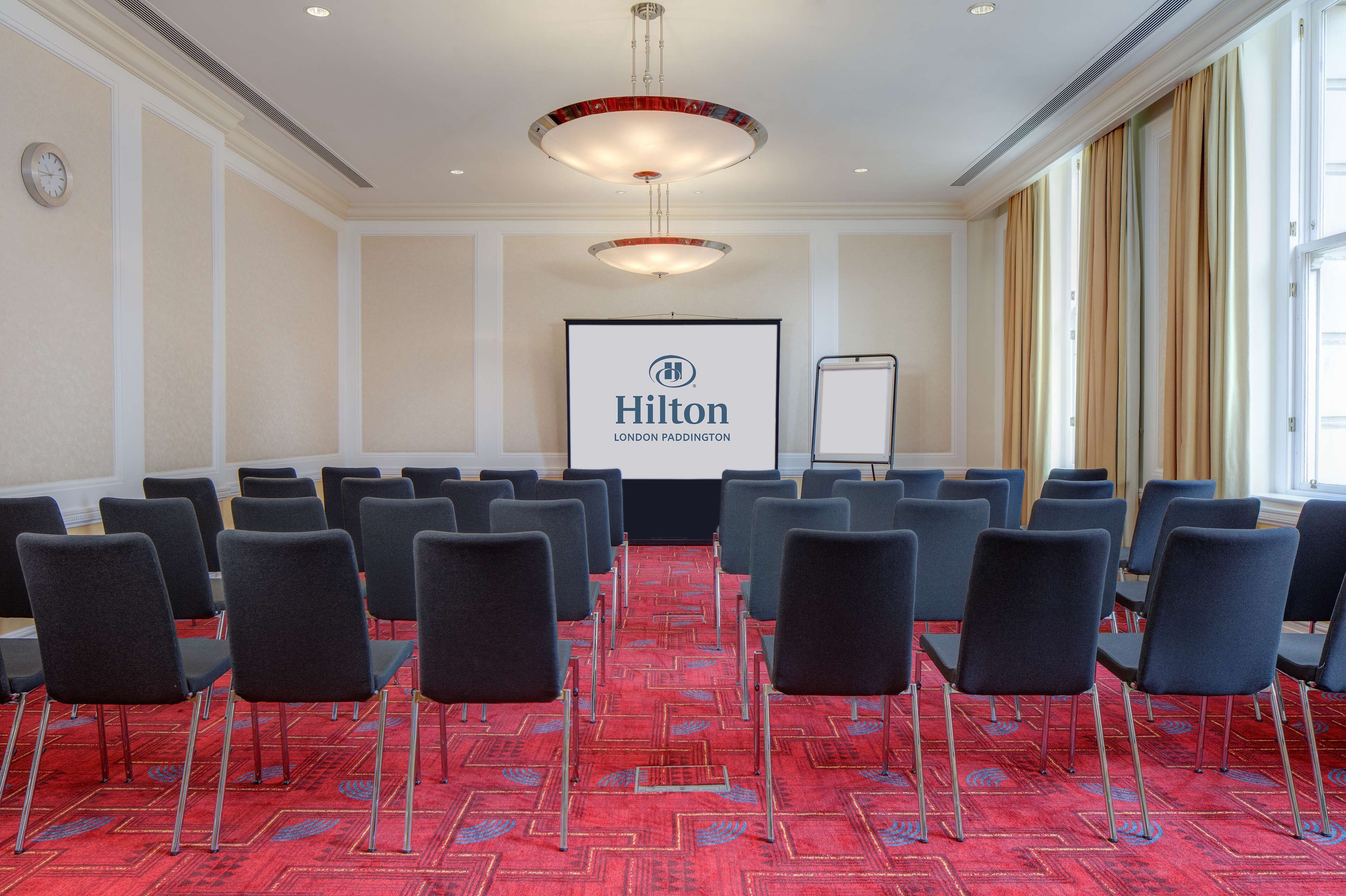 Ein Konferenzraum mit Reihen von Stühlen, einer Projektionsleinwand mit dem Logo von 'Hilton London Paddington' und einer Tafel.
