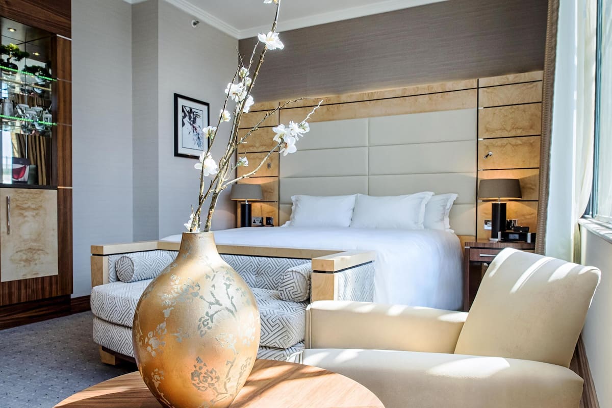 A hotel room with a bed, a sofa, a chair, a wooden table, and a vase with flowers.