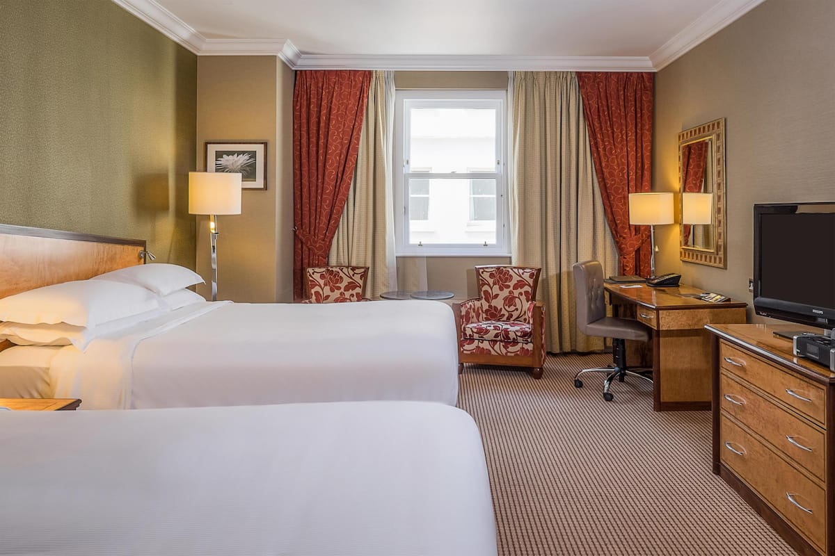 A hotel room with two beds, a desk, a chair, a TV, and a window with curtains.