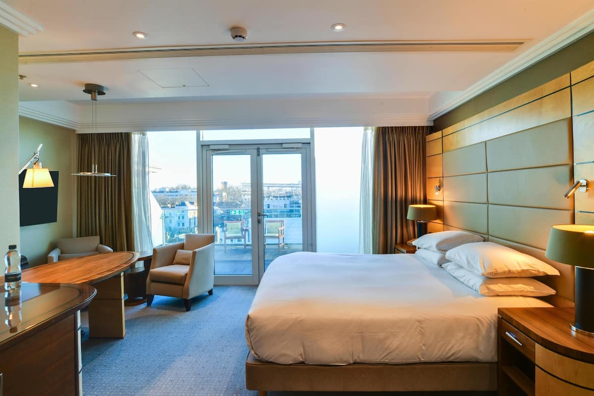 A hotel room with a large bed, a desk, a chair, a sofa, a view of the city, and a balcony.