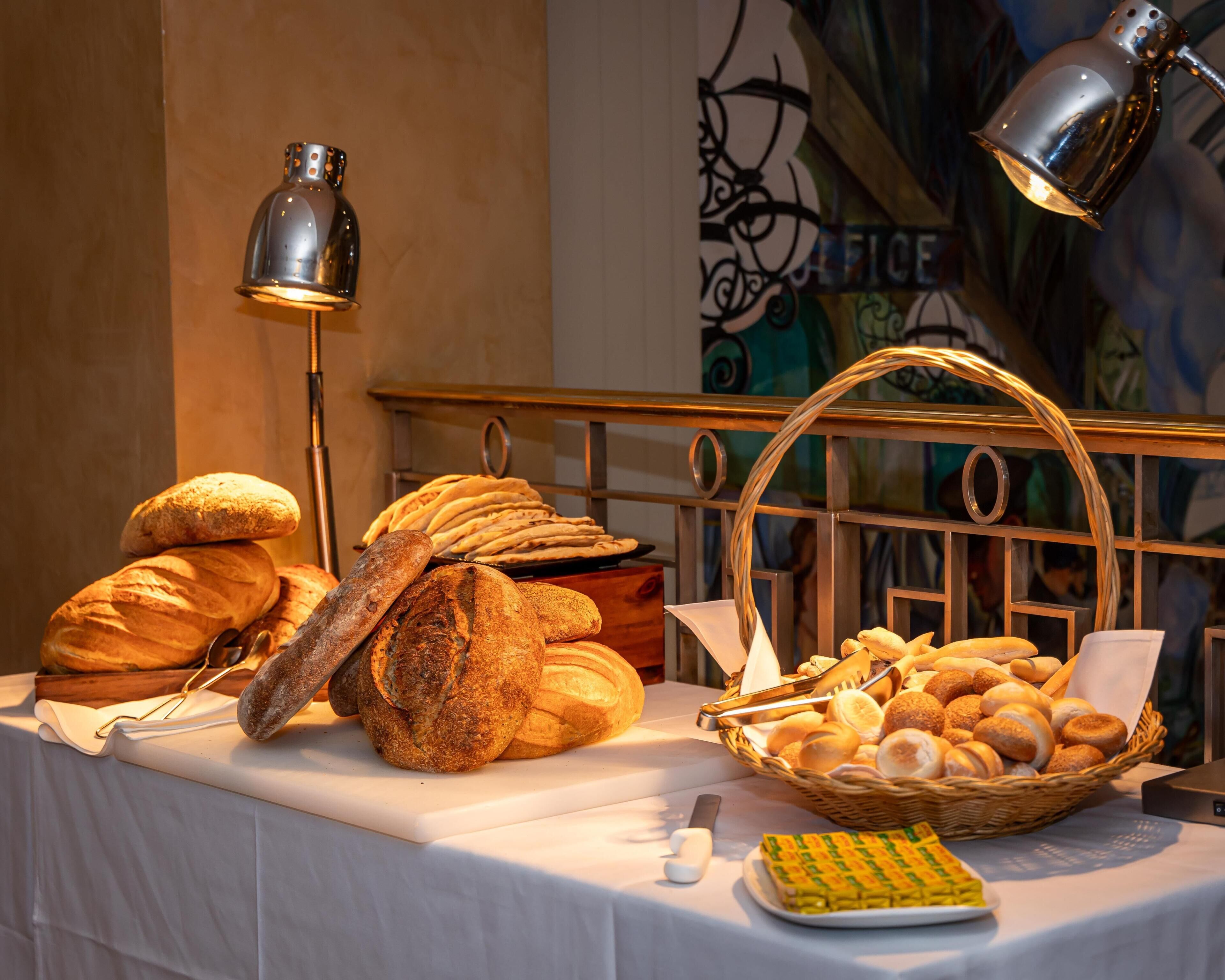 A table with various breads and pastries, a bread knife, and a basket of bread rolls.